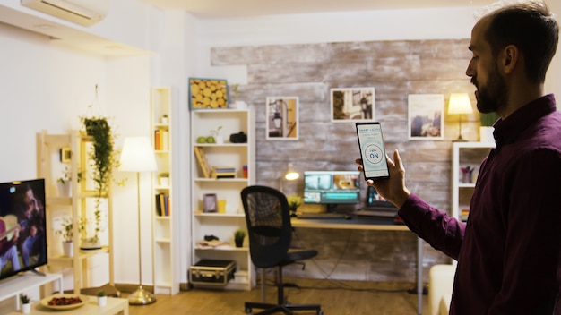 Mann gibt dem Smartphone einen Sprachbefehl, um das Licht im Haus einzuschalten und geht im Hintergrund zu seinem PC