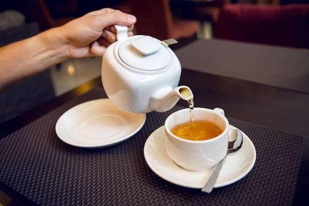 Mann gießt Tee aus einer weißen Teekanne in eine Teetasse, Untertasse und Teelöffel, eine Zuckerdose stehen auf dem Tisch