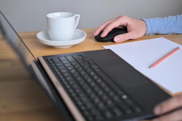 Mann hält Computermaus in der Hand und arbeitet am Laptop neben einer weißen Tasse Tee aus nächster Nähe