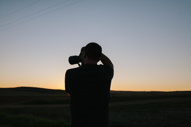 Foto mann hält eine kamera, während er während des sonnenuntergangs auf dem feld gegen den himmel steht