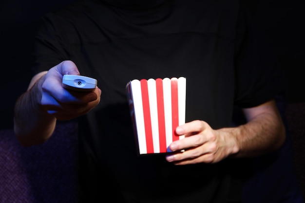 Mann hält eine TV-Fernbedienung und Popcorn in der Hand auf dunklem Hintergrund