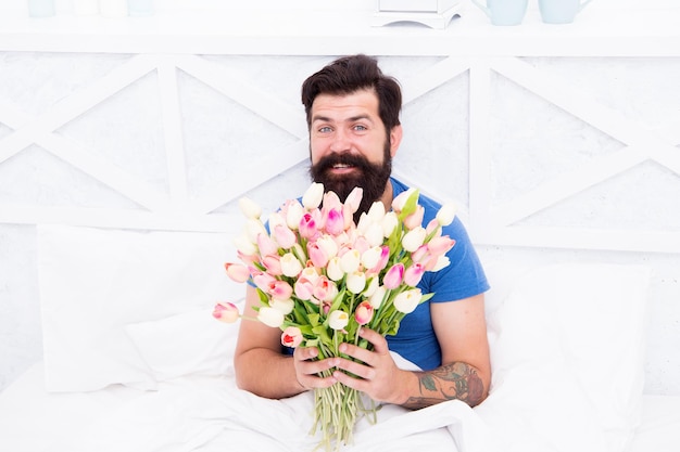 Mann hält Tulpenblumenstrauß beim Entspannen im Bett Zarte Blüte Frische Blumen überraschen Blumenlieferdienst Geburtstag Jubiläumsurlaub Geschenk für Ehepartner Bärtiger Hipster im Bett Frühling im Schlafzimmer