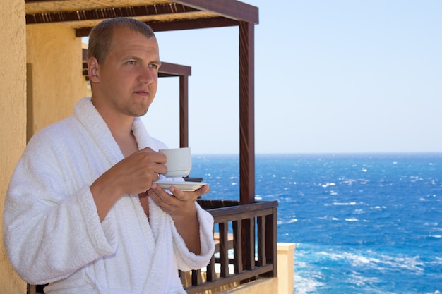 Mann im Bademantel mit Tasse Kaffee steht auf Balkon