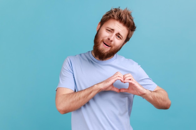 Mann im blauen T-Shirt, das die Hände in Form eines Herzens hält und eine romantische Geste der Liebesbeichte zeigt