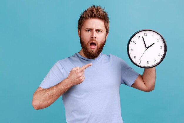 Mann im blauen T-Shirt zeigt mit verwirrtem Gesichtsausdruck auf die Deadline der Wanduhr