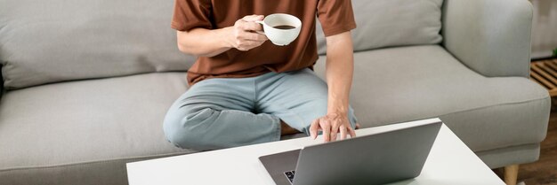 Mann im braunen T-Shirt, der mit einer Hand auf seinem Laptop tippt, während die andere Hand eine Tasse Kaffee hält