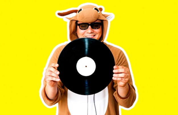 Mann im Cosplay-Kostüm einer Kuh mit Schallplatte isoliert auf gelbem Hintergrund. Kerl in der Tierpyjamanachtwäsche. Lustiges Foto mit Partyideen. Disco-Retro-Musik.