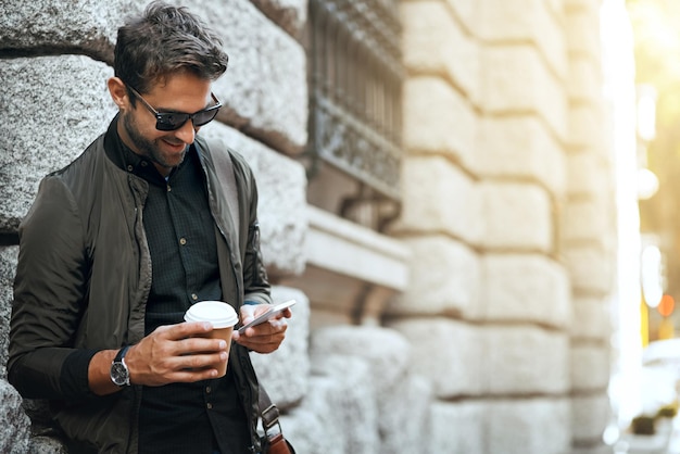 Mann im Freien und glücklich mit Smartphone an der Wand mit Kaffee in der Stadt für Social-Media-Post und Interaktion Mann Mitarbeiter und Lächeln oder zufrieden in New York mit Kommunikation und Networking