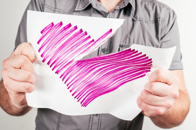 Mann im grauen Hemd reißt rosa Herz auf Blatt Papier in verschiedene Richtungen auf weißem Hintergrund gezeichnet. Gebrochene Liebe