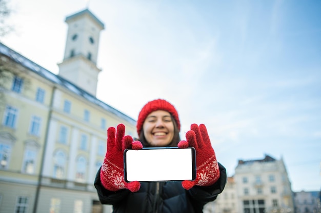 Mann im roten Winterhut, der Telefon mit weißem leerem europäischem Rathaus des leeren Bildschirms auf Hintergrund hält