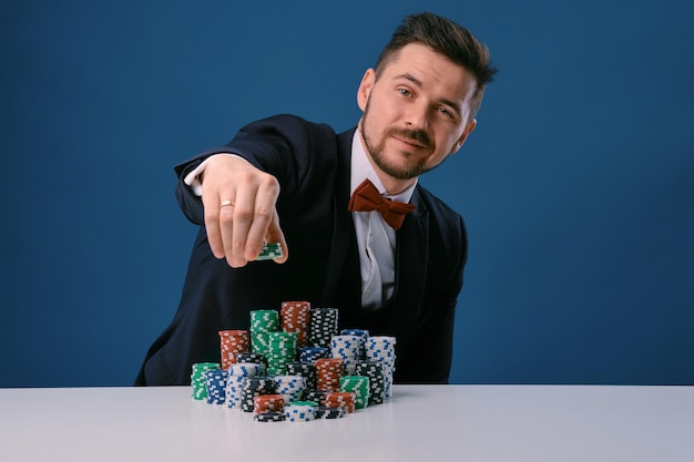 Mann im schwarzen Anzug sitzt an einem weißen Tisch mit farbigen Stapeln von Chips darauf und posiert auf blauem Studio...