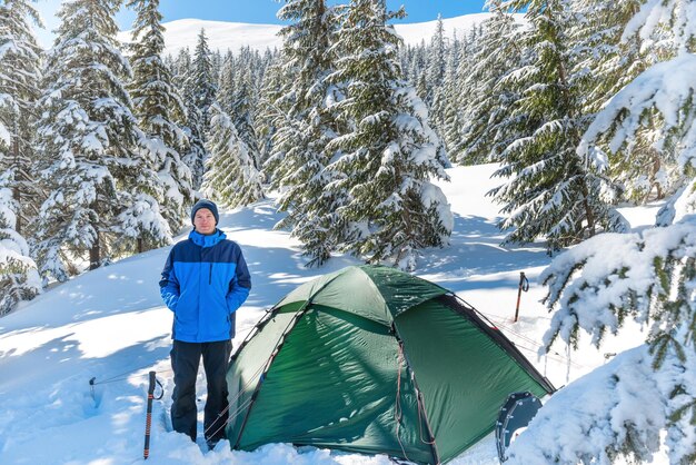 Mann im Wald in den Winterbergen mit tiefem Schnee, der in der Nähe des Zeltes steht und in die Kamera schaut