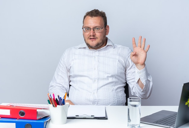 Mann im weißen Hemd mit Brille lächelnd und zeigt Nummer vier am Tisch sitzend mit Laptop-Büroordnern und Zwischenablage auf weiß