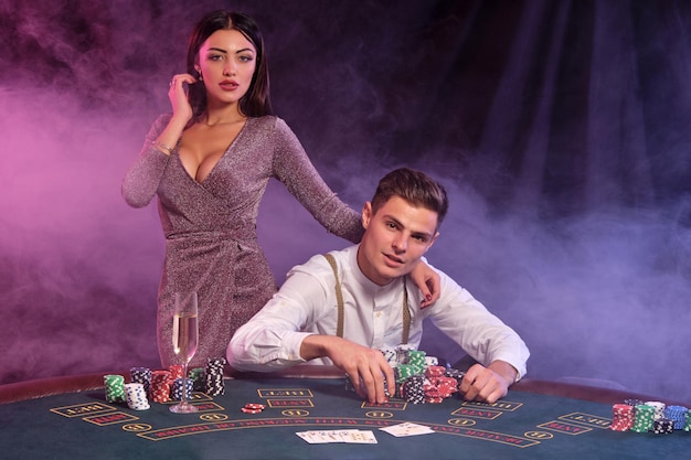 Mann im weißen Hemd spielt Poker im Casino und sitzt am Tisch mit Stapeln von Chips und Karten drauf Er feiert seinen Sieg mit Frau Schwarzer Rauch Hintergrund bunte Hintergrundbeleuchtung Glücksspiel Nahaufnahme