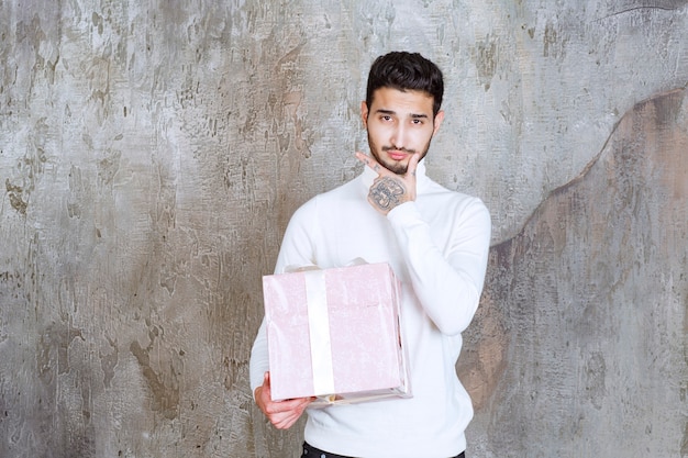 Foto mann im weißen pullover hält eine lila geschenkbox mit band umwickelt und sieht verwirrt oder nachdenklich aus.