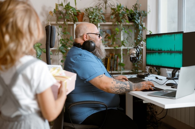 Mann in Übergröße mit Kopfhörern arbeitet am Computer, während Mädchen eine Geschenkbox im Zimmer hält