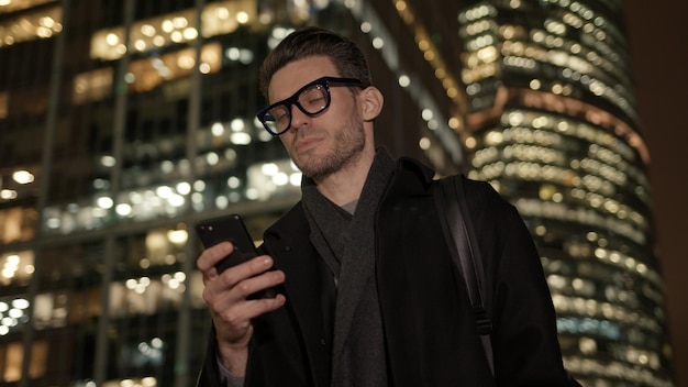 Mann in Brille mit Telefon auf dem Hintergrund von Geschäftsgebäuden in der Nacht