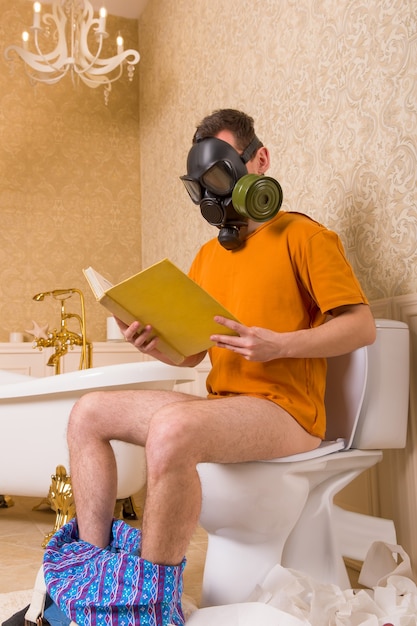 Mann in der Gasmaske, die auf Toilette sitzt und Buch liest