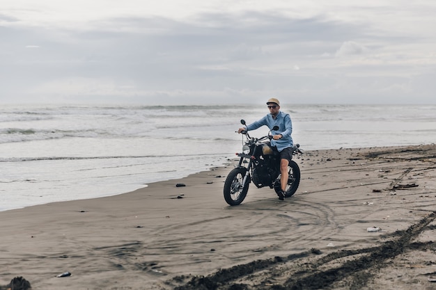 Mann in der Kappe, die Motorrad am Strand reitet. Moto Cross Dirtbiker am Strand Sonnenuntergang auf Bali. Junger Hipster-Mann, der Freiheit und aktiven Lebensstil genießt und Spaß auf einer Bikertour hat.
