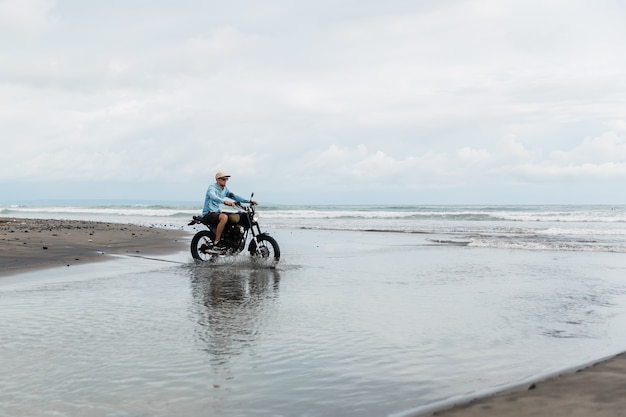 Mann in der Kappe, die Motorrad am Strand reitet. Moto Cross Dirtbiker am Strand Sonnenuntergang auf Bali. Junger Hipster-Mann, der Freiheit und aktiven Lebensstil genießt und Spaß auf einer Bikertour hat.