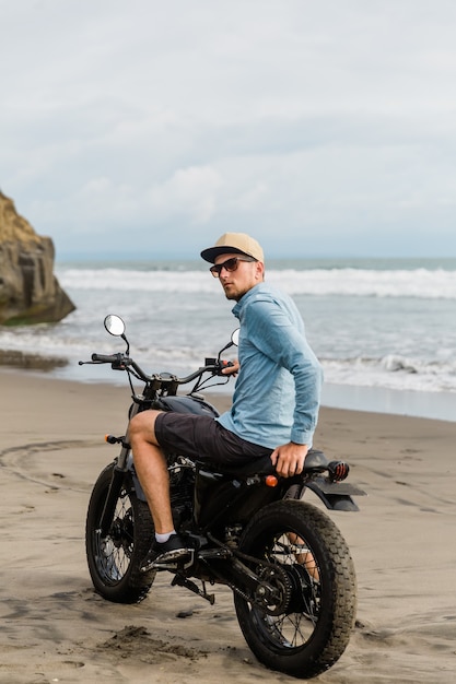 Mann in der Kappe, die Motorrad am Strand reitet. Moto Cross Dirtbiker am Strand Sonnenuntergang auf Bali. Junger Hipster-Mann, der Freiheit und aktiven Lebensstil genießt und Spaß auf einer Bikertour hat.