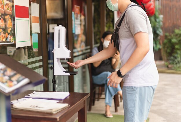 Mann in der medizinischen maske unter verwendung des automatischen alkoholgelspenders, der auf seine hand sprüht.