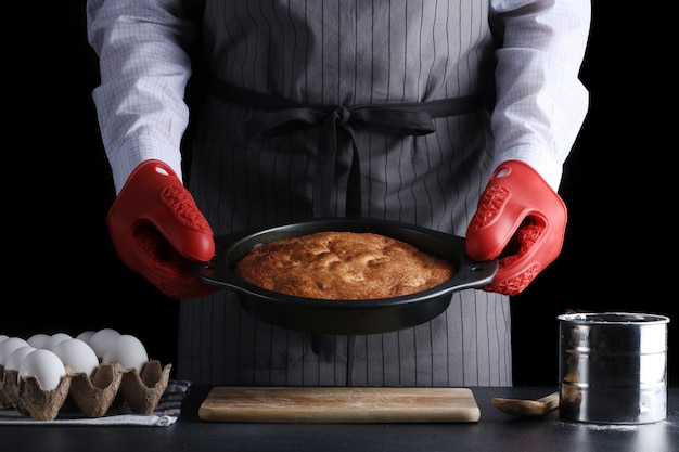 Mann in der Schürze, die frisch gebackenen Kuchen auf dunklem Hintergrund hält