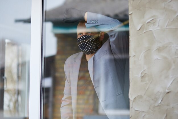 Mann in der schwarzen Maske und in den Medizinhandschuhen, die vom Fenster, Coronavirus-Quarantäne schauen