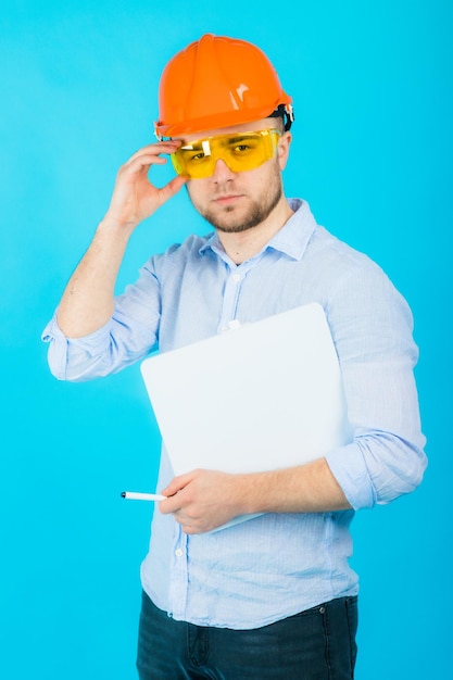 mann in einem blauen hemd in einem schützenden orangefarbenen helm und einem weißen notizbuch steht auf einem blauen hintergrund