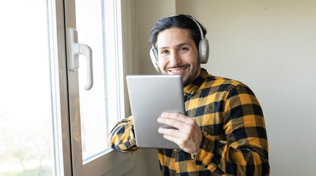 Mann in einem karierten Hemd, der sich mit einem Tablet in der Hand an ein Fenster lehnt, Musik hört und in die Kamera schaut