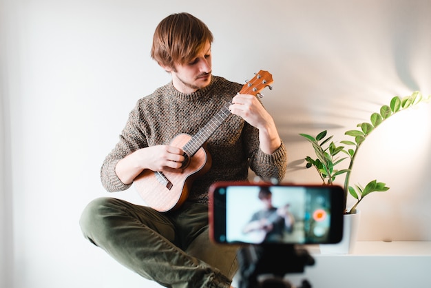 Mann in einem Pullover zieht den Vlog eines Spiels auf einer Ukulele zu Hause am Telefon aus.