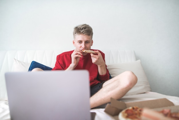 Mann in einem roten Sweatshirt sitzt zu Hause und isst ein Stück Pizza und schaut Filme auf einem Laptop