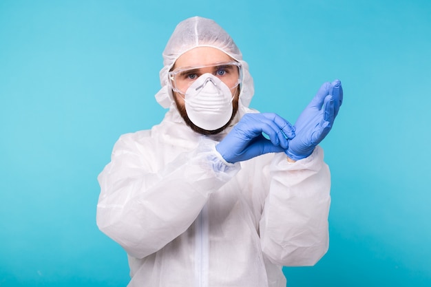 Mann in einem weißen Dekontaminationsanzug, der auf der blauen Wand isolierte medizinische Handschuhe anzieht