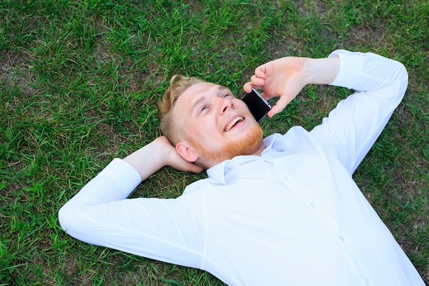 Mann in einem weißen Hemd liegt auf dem Gras und spricht an seinem Handy