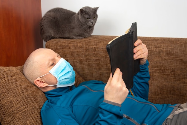 Mann in einer medizinischen Maske zu Hause in Quarantäne wegen einer Coronavirus-Epidemie liest eine Bibel, die auf einem Sofa neben einer grauen Katze sitzt