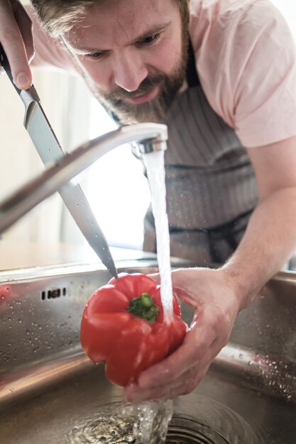 Foto mann in einer schürze hält pfeffer unter einen wasserstrahl, um ihn mit einem messer in der küche zu schneiden