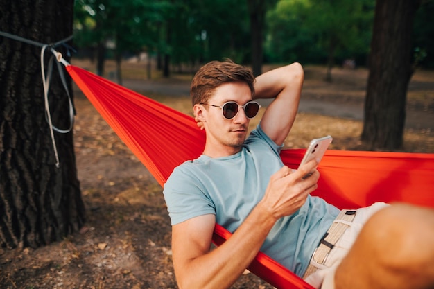 Mann in Freizeitkleidung, der sich im Wald ausruht, in einer Hängematte auf einem Baum liegt und ein Smartphone benutzt