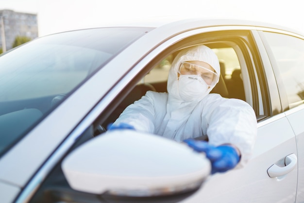 Mann in Hazmat-Anzug, Schutzhandschuhen und Schutzbrille beim Autofahren während einer Epidemie in der Quarantänestadt.