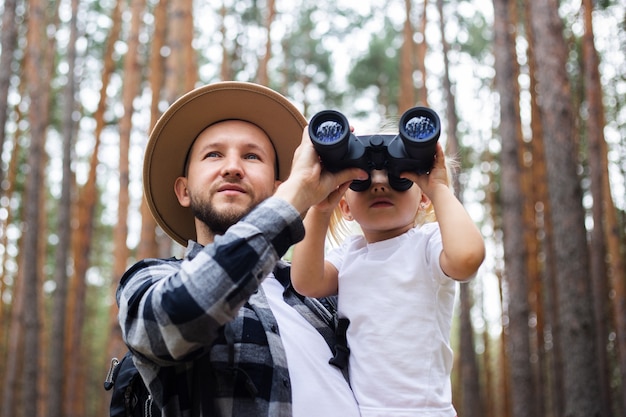 Mann in Hut und Rucksack und Kind schauen durch Fernglas beim Wandern im Wald. Familienwanderung in die Berge oder in den Wald.