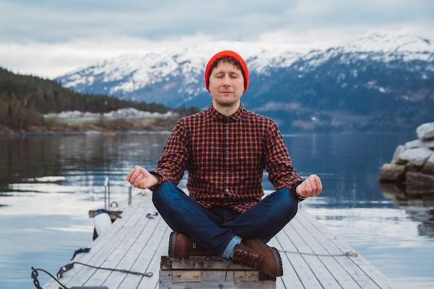 Mann in meditativer Position sitzt auf einem Holzsteg vor dem Hintergrund eines Berges und eines Sees