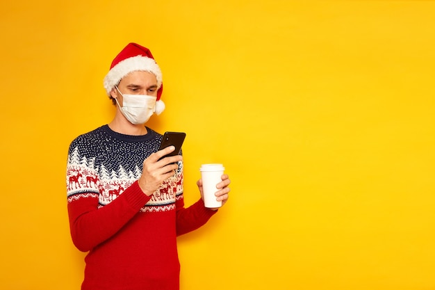 Mann in rotem Weihnachtsmütze Neujahrspullover mit Pappbecher Kaffee hält Telefon in den Händen liest Nachricht