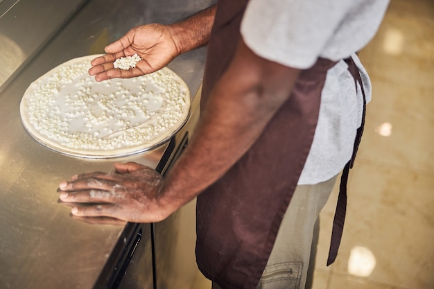 Mann in Schürze, der geriebenen Käse hält, während er ihn auf den Teig legt