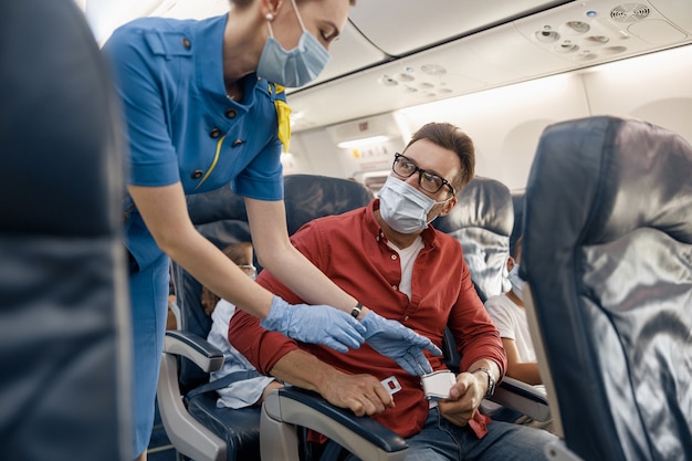 Mann in schützender Gesichtsmaske, der eine Flugbegleiterin ansieht, die ihm hilft, einen Sicherheitsgurt in einem Flugzeug für eine sichere Reise anzupassen und zu straffen. Reisen mit dem Flugzeug während der Covid19-Pandemie