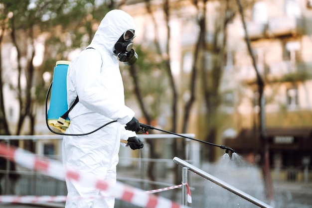 Mann in Schutzanzug und Maske sprüht Desinfektionsmittel auf Geländer im öffentlichen Raum Covid 19