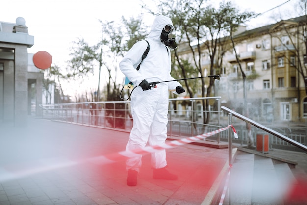 Mann in Schutzanzug und Maske sprüht Desinfektor auf das Geländer in der leeren öffentlichen Stelle im Morgengrauen in der Stadt der Quarantäne. Covid 19. Reinigungskonzept.