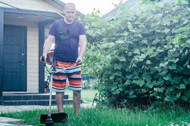 Mann in Schutzbrille, Shorts und T-Shirt mäht Rasen mit Benzintrimmer, vor dem Hintergrund von Weinberg und Eingang zum Haus