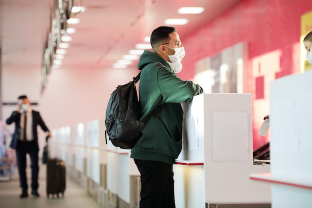 Mann in Schutzmaske mit Rucksack hinter dem Rücken, der an der Rezeption am Flughafen steht