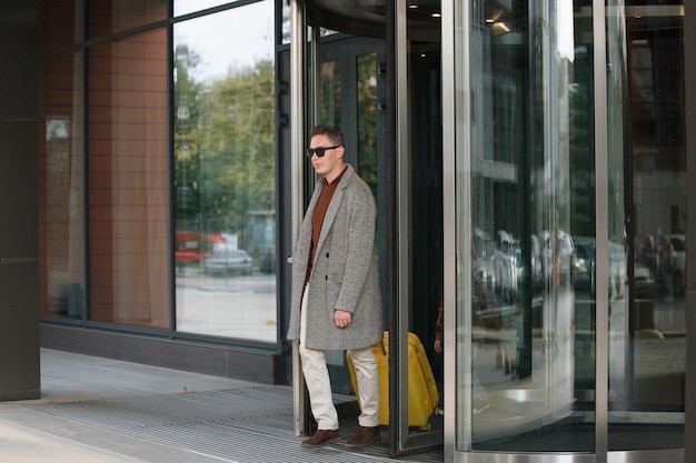 Mann in schwarzer Brille, mit gelbem Koffer in beiger Hose, grauem Mantel, verlässt das Hotel.