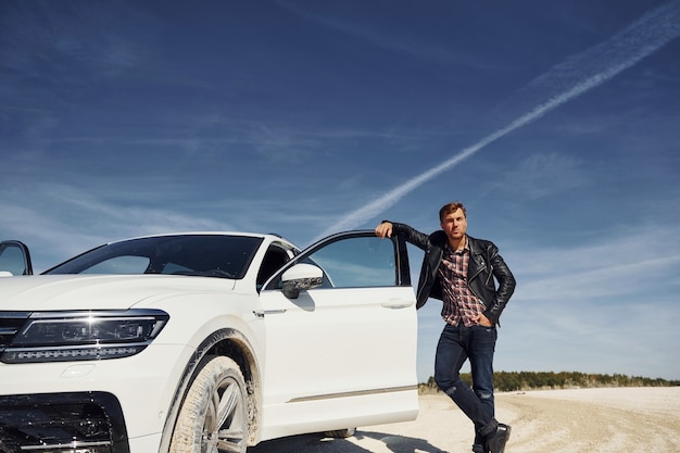 Mann in schwarzer Lederjacke steht in der Nähe seines geparkten weißen Autos im Freien vor blauem Himmel.