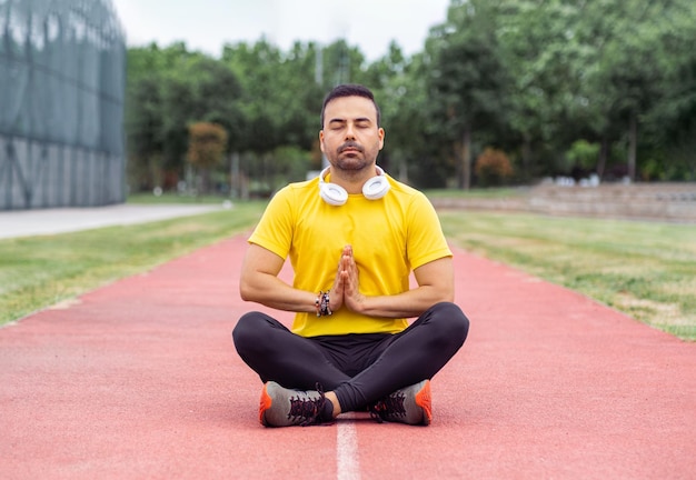 Mann in sportlicher Kleidung meditiert im Lotussitz auf der Laufstrecke im städtischen Stadion im Freien